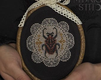 May include: An oval cross-stitch embroidery featuring a red and brown beetle design, surrounded by a white floral pattern, set against a black background. The embroidery is framed in a wooden hoop and adorned with a cream-colored lace bow. The text "MIDNIGHT STITCH" is visible.