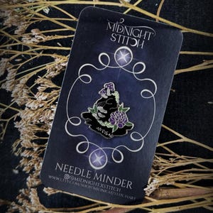 Peut inclure: Un aimant à aiguilles en émail noir représentant un champignon violet et une fleur sur une carte rectangulaire avec le texte Midnight Stitch et Needle Minder en blanc, entouré d'une bordure décorative en ligne blanche.