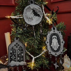 May include: Cross-stitched Christmas ornaments in black and white hang on a small tree. The ornaments feature a bat, a ghost, and a gothic window design. Each ornament has a black ribbon for hanging and decorative charms.