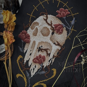 Puede incluir: Arte de punto de cruz sobre tela negra con un diseño de calavera con detalles florales. La calavera es blanquecina con sombreado marrón, rodeada de flores rojas y un halo dorado. El texto "Midnight Stitch" es visible.