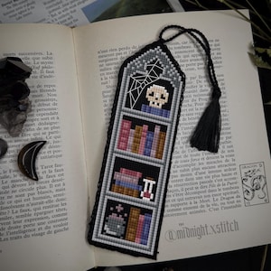 Könnte beinhalten: Schwarz-grauer Kreuzstich-Lesezeichen mit einem Totenkopf, Spinnennetz und Bücherdesign. Das Lesezeichen hat eine schwarze Quaste und wird auf einem offenen Buch mit Text gezeigt.