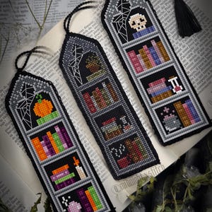 Könnte beinhalten: Drei handgefertigte, schwarze Kreuzstich-Lesezeichen mit Halloween-Motiven. Jedes Lesezeichen zeigt ein anderes Design: einen Kürbis, ein Bücherregal und einen Totenkopf. Die Lesezeichen haben schwarze Quasten.