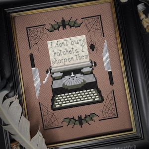 Op de afbeelding: Kruissteekkunstwerk in een zwarte lijst met een vintage typemachine met de tekst "I don't bury hatchets, I sharpen them". Het ontwerp bevat vleermuizen, spinnenwebben en messen, met een bruine achtergrond. Het kunstwerk heeft een gotisch thema.