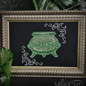 May include: A cross-stitch embroidery featuring a green cauldron on a black background, framed in a metallic gold-toned ornate frame. The design includes white decorative scrollwork in the corners and the text Midnight Stitch in the bottom right.