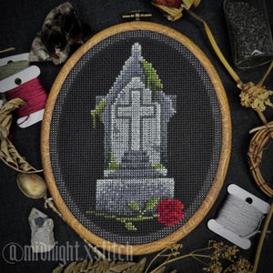 Puede incluir: Una lápida en forma de cruz con una rosa roja y enredaderas verdes, bordada en hilo negro, gris y blanco sobre tela negra, enmarcada en un aro de madera.