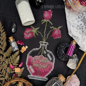 May include: A black cross-stitch design featuring a vase of pink roses. The image includes small glass bottles with cork stoppers, crystals, dried rose petals, and the text "@midnight.xstitch".