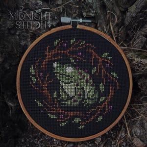 Pode incluir: Um bastidor de bordado circular de madeira contendo um desenho em ponto cruz sobre tecido preto. O design apresenta um sapo verde sentado dentro de uma coroa circular de ramos castanhos com pequenas folhas verdes e toques rosa. O texto MIDNIGHT STITCH é visível.