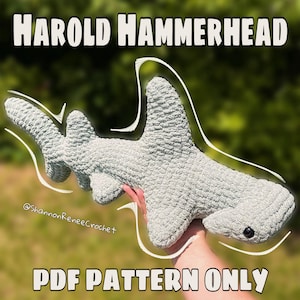 May include: A crocheted hammerhead shark in light gray, held up against a blurred green background. The shark has a textured surface, black eyes, and the text "HAROLD HAMMERHEAD" and "PDF PATTERN ONLY" in white. The artist's name is also visible.