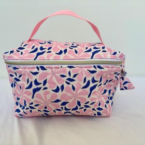 May include: Pink floral print cosmetic bag with a matching pink handle. The bag features a white background with pink flowers and blue leaf accents. It has a gold zipper and a small fabric tag attached to the zipper.