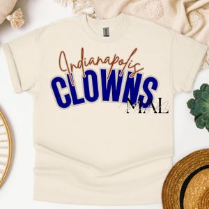 May include: Cream-colored t-shirt featuring the text "Indianapolis CLOWNS" in blue and red lettering. The shirt has short sleeves and a crew neck. The design includes a shadow effect, enhancing the text's visibility.