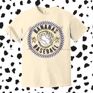 May include: Cream-colored t-shirt featuring a baseball-themed graphic. The design includes a baseball, glove, and the words "BANANAS BASEBALL" in a circular arrangement. Two cartoon banana characters are on either side of the baseball.