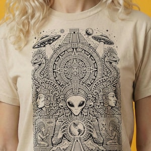 May include: Beige t-shirt with a black and white graphic depicting an alien holding a globe, surrounded by Mayan-inspired imagery, pyramids, and UFOs. The shirt has short sleeves and a crew neck.
