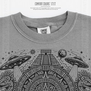 Puede incluir: Una camiseta gris Comfort Colors 1717 con un diseño gráfico de ovnis, una pirámide y elementos celestiales. La camiseta tiene cuello redondo y mangas cortas. El texto "COMFORT COLORS 1717" está impreso en la parte superior.