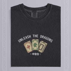 May include: A charcoal gray crew neck t-shirt featuring a graphic of three mahjong tiles. Above the tiles, the text reads "UNLEASH THE DRAGONS" in a white arched font. Below the tiles, the text "中發白" is printed in white with small diamond accents.