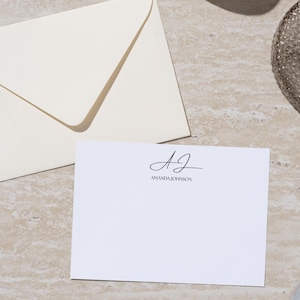 Pode incluir: Conjunto de papelaria marfim com um envelope dobrado e um cartão em branco com as iniciais "AJ" e o nome "Amanda Johnson" impressos em escrita preta. Uma caneta prateada está ao lado. O conjunto está disposto sobre uma superfície clara e texturizada.