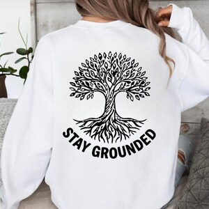 May include: A white long-sleeve sweatshirt featuring a black graphic on the back. The graphic depicts a detailed tree with visible roots and branches, accompanied by the text "STAY GROUNDED" printed in a bold, sans-serif font below the tree.