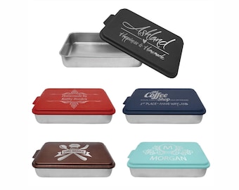 May include: A collection of rectangular baking pans with lids. The lids are in various colors, including black, red, blue, brown, and teal. Each lid has custom text, such as "Ashland" and "Homemade by Kathy Bozden."