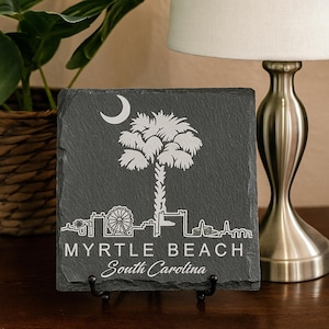 May include: A square, dark gray slate coaster featuring a white etched design of a palm tree, crescent moon, and the Myrtle Beach, South Carolina skyline. The text "MYRTLE BEACH South Carolina" is below the skyline.
