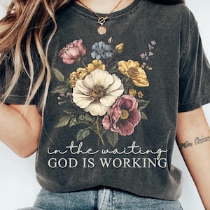 May include: Dark gray t-shirt featuring a floral bouquet in shades of pink, yellow, white, and blue. The text "in the waiting GOD IS WORKING" is printed below the flowers. The shirt has a vintage, washed look.