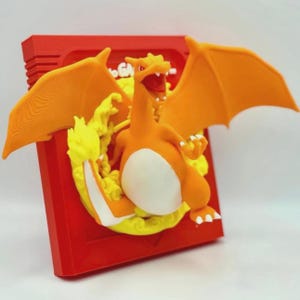 May include: A 3D printed figurine of a Charizard Pokemon, a fire-type Pokemon, in a red and yellow Game Boy cartridge frame. The Charizard is orange with white underbelly and yellow flames.
