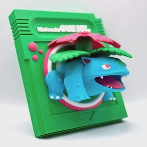 May include: A 3D printed figurine of a blue Pokemon character, Venusaur, with a green leafy head and pink flower, emerging from a green Nintendo Game Boy cartridge.