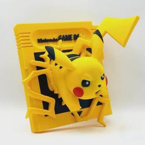 May include: A yellow 3D printed figurine of Pikachu, a popular Pokemon character, emerging from a yellow Nintendo Game Boy cartridge. The figurine has black eyes and red cheeks.