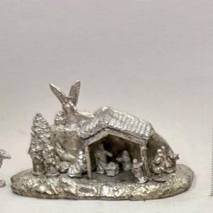 May include: A miniature silver-colored nativity scene with a camel figurine. The scene includes a stable, figures, and an angel. The base is approximately 2 inches long.