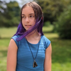 May include: A young person with purple hair wearing a blue striped dress and a necklace with a dark brown pendant.