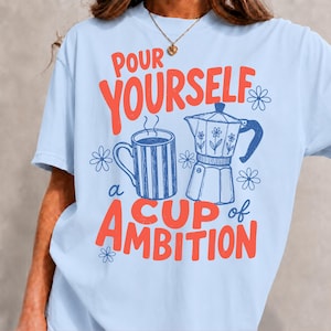 K&ouml;nnte beinhalten: Ein hellblaues T-Shirt mit einem Grafikdruck, der den Text "POUR YOURSELF a CUP of AMBITION" in Rot zeigt. Das Design umfasst eine blau gestreifte Kaffeetasse, eine blaue Espressokanne mit Blumenmotiven und mehrere kleine blaue Blumen-Icons.