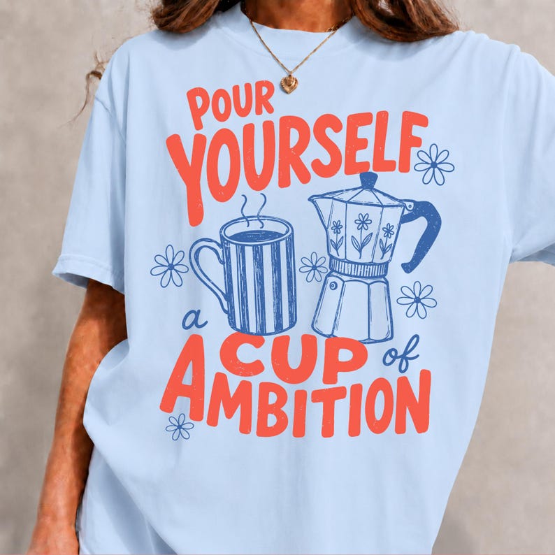 K&ouml;nnte beinhalten: Ein hellblaues T-Shirt mit einem Grafikdruck, der den Text "POUR YOURSELF a CUP of AMBITION" in Rot zeigt. Das Design umfasst eine blau gestreifte Kaffeetasse, eine blaue Espressokanne mit Blumenmotiven und mehrere kleine blaue Blumen-Icons.