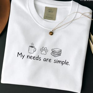 Puede incluir: Camiseta blanca de cuello redondo con un gráfico negro de una taza de café, una huella de pata y libros. El texto "My needs are simple" está debajo. Una pequeña etiqueta y un collar son visibles.