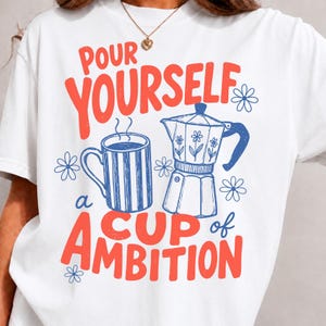 K&ouml;nnte beinhalten: Ein wei&szlig;es T-Shirt mit einem Grafikdruck, der den roten Schriftzug "POUR YOURSELF A CUP OF AMBITION" sowie blaue Strichzeichnungen einer gestreiften Kaffeetasse und einer Espressokanne zeigt, erg&auml;nzt durch kleine blaue Blumenmotive.