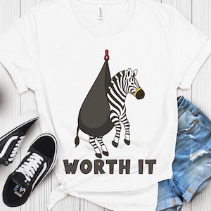 May include: White t-shirt featuring a cartoon zebra with a black and white striped pattern, wearing a yellow nose ring, and hanging from a black teardrop-shaped object. The text "WORTH IT" is printed in bold, black letters below the zebra.
