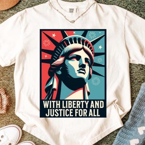 May include: White t-shirt featuring a graphic of the Statue of Liberty in red, blue, and white. The design includes the text "WITH LIBERTY AND JUSTICE FOR ALL" in a bold, black font. The shirt is made of a soft material.