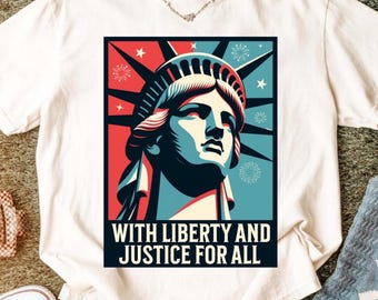 May include: White t-shirt featuring a graphic of the Statue of Liberty in red, blue, and white. The design includes the text "WITH LIBERTY AND JUSTICE FOR ALL" in a bold, black font. The shirt is made of a soft material.