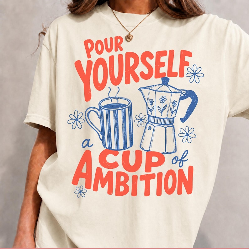 K&ouml;nnte beinhalten: Ein cremefarbenes T-Shirt mit einem Grafikdruck, der den roten Schriftzug "POUR YOURSELF a CUP of AMBITION" zeigt, umgeben von einer blau gestreiften Kaffeetasse und einer blauen Espressokanne, akzentuiert durch kleine blaue Blumenmotive.