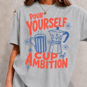 K&ouml;nnte beinhalten: Ein hellgraues T-Shirt mit einem Grafikdruck, der den Text "POUR YOURSELF a CUP of AMBITION" in Rot zeigt. Das Design umfasst eine blau gestreifte Tasse und eine blaue Mokkakanne, die beide von kleinen blauen Blumenillustrationen umgeben sind.