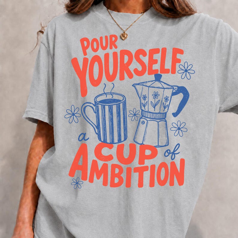 K&ouml;nnte beinhalten: Ein hellgraues T-Shirt mit einem Grafikdruck, der den Text "POUR YOURSELF a CUP of AMBITION" in Rot zeigt. Das Design umfasst eine blau gestreifte Tasse und eine blaue Mokkakanne, die beide von kleinen blauen Blumenillustrationen umgeben sind.
