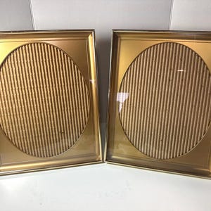 May include: A pair of rectangular gold-toned picture frames featuring a central oval cutout. Each frame contains a textured, vertically ribbed tan cardboard insert behind a clear glass pane, creating a layered appearance within the metallic border.