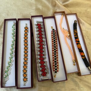 May include: A collection of beaded bracelets in various colors and designs, displayed in rectangular boxes. The bracelets feature patterns of green, orange, red, black, and peach beads. Some have silver or gold-toned clasps.