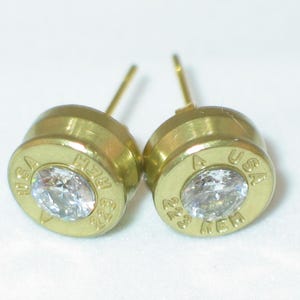 May include: A pair of gold-colored stud earrings. Each earring features a clear, round gemstone set in a casing with the text "USA REM" engraved on it. The earrings are on a white background.