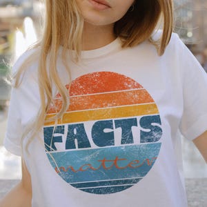 May include: White t-shirt featuring a retro-style circular design with horizontal stripes in orange, yellow, and blue. The word "FACTS" is printed in bold, blue letters, with "matter" in a smaller, cursive font.