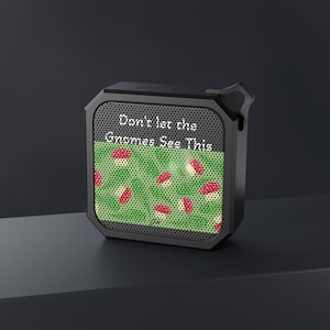 May include: Black, square portable speaker with a mesh front. The speaker's screen displays a green field with red and white mushrooms. Text on the screen reads, "Don't let the Gnomes See This."