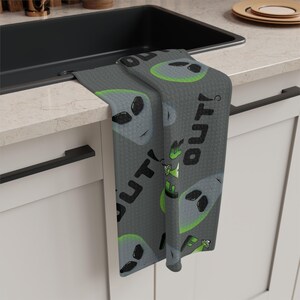 May include: Gray kitchen towel featuring a repeating pattern of alien faces with black eyes and green accents. The towel also has the text "OUT!" in black. The towel is hanging over a sink.