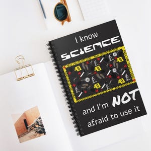 May include: Black spiral-bound notebook with the text "I know SCIENCE and I'm NOT afraid to use it." The cover features a crime scene design with evidence markers, footprints, and knives. The notebook is open on a white surface.