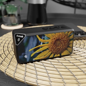 May include: Black rectangular Bluetooth speaker with a sunflower design on the front. The speaker has three buttons on top and a Bluetooth logo on the side. The speaker is on a woven mat.