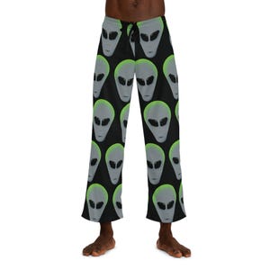 Op de afbeelding: Zwarte pyjamabroek met een herhaald patroon van grijze aliengezichten met groene accenten. De broek heeft een taille met trekkoord en een relaxte pasvorm. De aliengezichten zijn gedetailleerd met grote zwarte ogen en een gladde, grijze huidskleur.