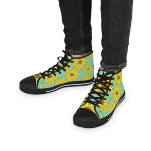 May include: High-top canvas trainers with a black sole and laces. The trainers feature a repeating pattern of yellow sunflowers on a light teal background. The shoes are worn with black denim jeans.