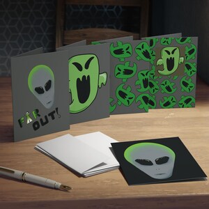 Peut inclure: Cartes de vœux avec des motifs d'extraterrestres et de fantômes. Les cartes sont grises avec des illustrations vertes et noires. Une carte porte l'inscription "FAR OUT!". Un stylo et des enveloppes blanches sont également présents.