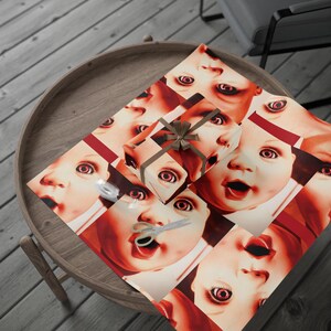 May include: A wrapped present with a brown bow rests on wrapping paper patterned with doll faces. The paper is red and white, with scissors and tape. The gift is on a wooden table.